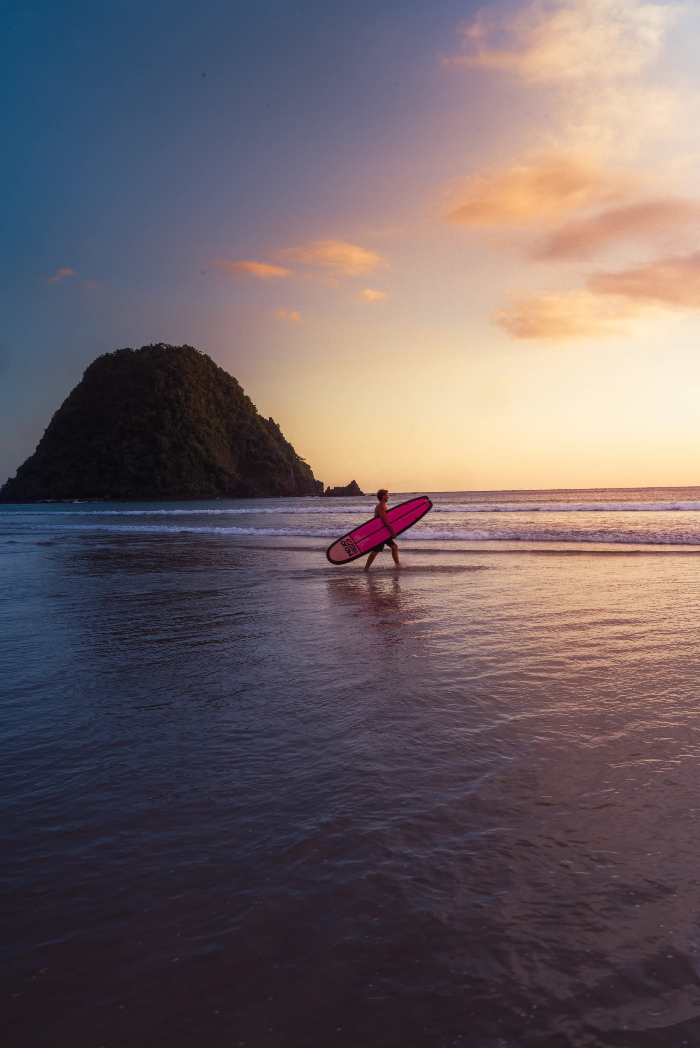 A surfer carrying a board into the Pulau Merah sunset